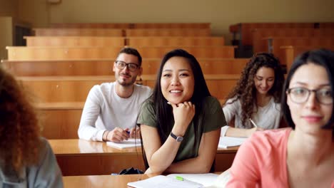 Pretty-Asian-girl-is-raising-hand-and-asking-question-during-lecture-at-college,-her-classmates-are-sitting-at-desks-smiling-and-laughing.-Education-and-youth-concept.