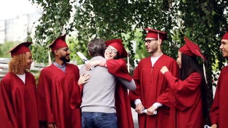 Lenta-de-profesor-universitario-abrazando-a-sus-estudiantes-y-agitando-las-manos-en-el-día-de-la-graduación-con-alegría-y-orgullo.-Árboles-verdes,-edificio-de-la-institución-educativa-es-visible.