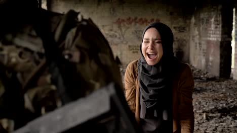 Young-muslim-woman-in-hijab-screaming-and-crying-behind-soldier-with-ammunition-and-weapon,-standing-in-abandoned-building,-war-concept