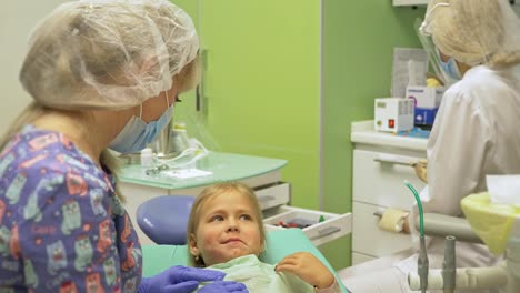 Niño-con-una-madre-en-la-recepción-de-un-dentista.-La-chica-se-encuentra-en-la-silla,-detrás-de-su-madre.-El-médico-trabaja-con-un-asistente.-Procedimiento-para-la-perforación-de-un-diente.-Establecer-un-sello-temporal.