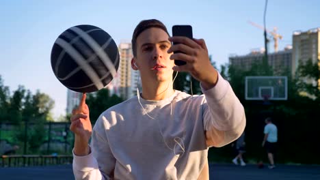 Hübscher-Mann-Spinnen-Basketball-am-Finger-und-nehmen-Selfie-oder-mit-video-Chat,-tragen-Kopfhörer,-lächelnd,-Park-im-Hintergrund