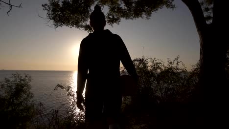 A-silhouette-of-a-basketball-player-girl-comes-with-a-ball-in-her-hand,-coming-up-to-the-slope-with-trees-around.-Looks-at-the-sun-shining-over-the-sea.-Morning-dusk.-Backside-view