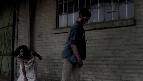 Two-zombies-are-walking-with-an-abandoned-house-on-the-background.-Brunette-girl-with-wounded-face-and-bloody-white-dress-and-wounded-male-zombie-are-walking-outdoors.-Accelerating-footage