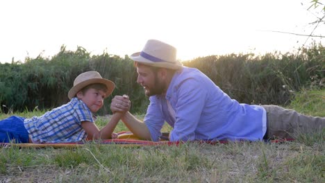 happy-father-and-son-kid-play-together-in-arm-wrestling-lying-on-plaid-outdoors-on-background-of-sunset-in-rural