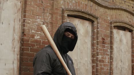 Street-hooligan-in-black-mask-with-baseball-bat-on-criminal-street-district-looking-to-camera