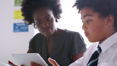 Close-Up-Of-Female-High-School-Tutor-With-Male-Student-Wearing-Uniform-Using-Digital-Tablets-For-One-To-One-Tuition-In-Classroom