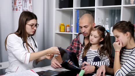 Father-with-two-daughters-on-consultation-in-the-office-of-the-family-doctor.-Female-doctor-talking-with-the-father-of-two-daughters,-and-shows-him-x-ray.-youngest-daughter.