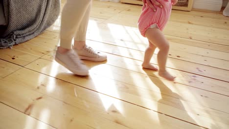 Tracking-right-shot-of-mother-supporting-her-barefoot-baby-daughter-in-pink-bodysuit-learning-to-walk-on-hardwood-floor-at-home