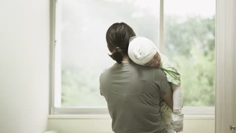 Madre-asiática-estaba-sosteniendo-una-niña-enferma-en-su-hombro-junto-a-la-ventana-del-hospital,-niña-está-enferma-con-dermatitis-atópica.-Estaba-envuelta-con-un-vendaje-en-los-brazos-y-las-piernas-del-cuello-de-la-cabeza.-Real-live