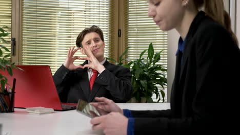 A-young-guy-in-a-suit-shows-a-heart-sign-to-a-female-employee.-Work-in-the-office,-humor-concept.-Work-in-office.-60-fps