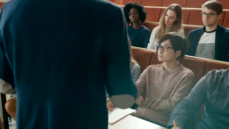 Cámara-frente-a-la-clase:-Vista-posterior-de-la-Universidad-profesor-da-lectura-a-un-aula-llena-de-estudiantes-Multi-étnica.-Profesor-Gesticulates-con-sus-manos-mientras-los-estudiantes-escuchar.