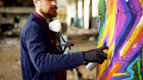 Bearded-man-graffiti-painter-is-using-aerosol-paint-to-decorate-pillar-in-old-industrial-building.-Modern-urban-art,-creative-young-people-and-hobby-concept.