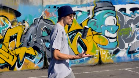 Young-man-is-walking-along-the-street-with-graffiti-painting-on-the-background-and-a-spray-in-his-hands