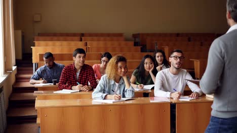 Grupo-multiétnico-de-estudiantes-es-respondiendo-a-preguntas-del-profesor,-jóvenes-inteligentes-están-hablando-y-sonriendo-mientras-el-maestro-les-pide-comprobación-de-conocimientos.