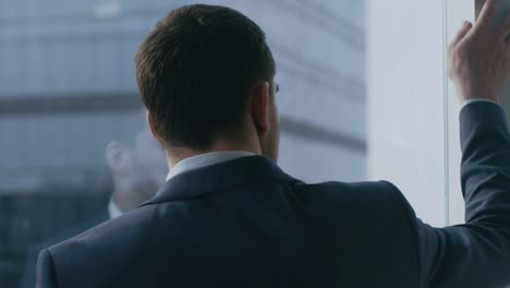 Close-up-Shot-of-the-Thoughtful-Businessman-in-a-Suit-Standing-in-His-Office-and-Contemplating-Next-Big-Business-Deal,-Looking-out-of-the-Window.-Big-City-Business-District-Panoramic-Window-View.