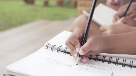 Alumno-asiático-dibujar-en-los-cuadernos-de-rodillas-durante-una-clase-al-aire-libre