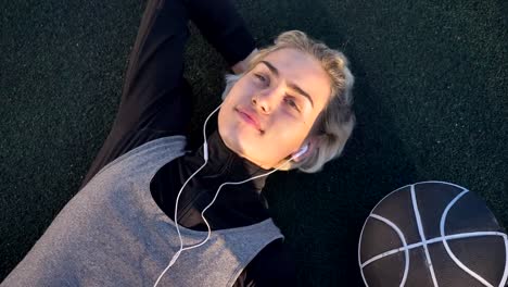 Dreamy-young-female-basketball-player-lying-on-court-and-listening-music-through-earphones,-ball-placing-near-her,-peaceful-and-calm