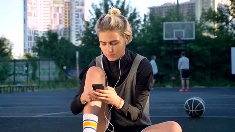 Pensive-young-beautiful-woman-sitting-on-basketball-court-and-typing-on-phone,-wearing-earphones,-men-playing-in-background