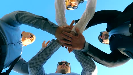 Young-business-partners-standing-and-stacked-hands-together-outdoor.-Successful-colleagues-putting-their-arms-in-centre-and-raising-them-up-to-blue-sky.-Unity-and-teamwork-concept.-Low-angle-view