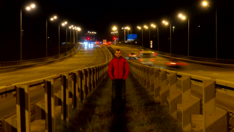 Ein-Mann-steht-auf-der-Autobahn.-Autobahn-Verkehr-verschwommen-Autos-bei-Nacht-Zeitraffer.-Autos-bewegen-unterwegs-auf-Brücke-Abend-Zeitraffer.-4K-UHD