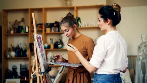 Estudiante-y-profesor-de-arte-de-las-mujeres-jóvenes-están-pintando-juntos,-hablando-y-sonriendo-durante-la-clase-en-estudio-creativo.-Bellas-Artes,-educación-y-concepto-de-la-gente.