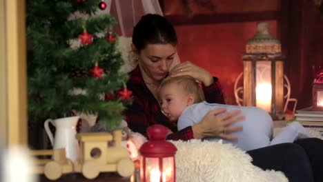 Mother-breastfeeding-her-toddler-son-sitting-in-cozy-armchair-near-Christmas-tree,-wintertime