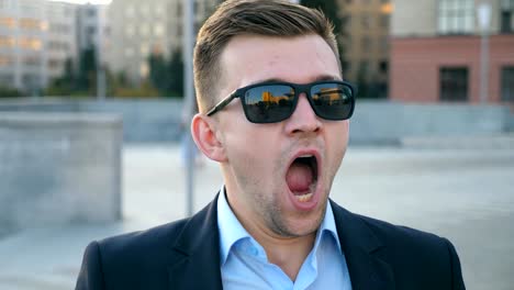 Portrait-of-tired-businessman-in-sunglasses-yawning-outdoor.-Exhausted-sleepy-man-opening-his-mouth-outside.-Angry-guy-yelling-to-camera.-Handsome-male-entrepreneur-screaming-at-city-street.-Close-up-Slow-motion