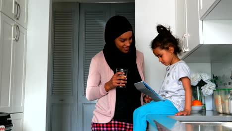 Joven-madre-con-un-hijab-bebiendo-un-vaso-de-agua-cerca-de-su-hija-en-la-cocina-4k