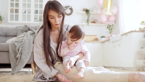 Beautiful-young-mother-sitting-on-floor-at-home-and-playing-with-baby-daughter-on-lap-patting-gently-her-tiny-feet