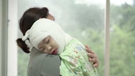 Asian-mother-was-holding-a-sick-baby-girl-at-her-shoulder-beside-the-window-in-hospital,-Girl-are-sick-with-Atopic-dermatitis.-She-was-wrapped-with-a-bandage-on-the-head-neck-arms-and-legs.-Real-live