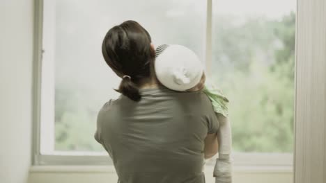 Asian-mother-was-holding-a-sick-baby-girl-at-her-shoulder-beside-the-window-in-hospital,-Girl-are-sick-with-Atopic-dermatitis.-She-was-wrapped-with-a-bandage-on-the-head-neck-arms-and-legs.-Real-live