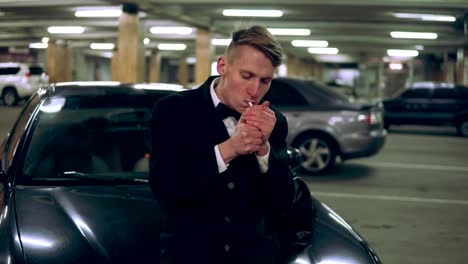 Young-man-in-a-black-suit-with-a-bow-tie-smoking-a-cigarette-sitting-at-the-bonnet-of-the-black-car-in-the-parking.-Waiting-for-someone-and-holding-a-big-bag.-Getting-up-and-going-away.-Front-view