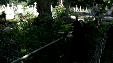 Pained-funeral-woman-widow-placing-a-crown-on-old-grave-with-statue-covered-in-moss-ivy-and-weeds-crying-down-her-knees