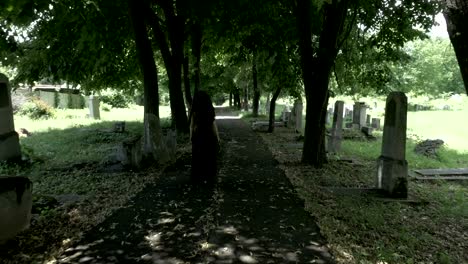 Funeral-gothic-widow-woman-in-black-holding-a-crown-in-hand-walking-in-the-old-cemetery-alley-filled-with-fallen-tree-leaves