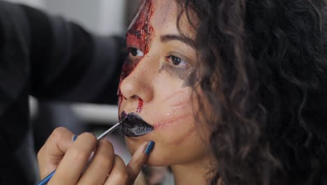 Make-up-artist-make-the-girl-halloween-make-upin-studio.Halloween-face-art.-Woman-applies-on-black-lipstick-with-brush-on-lips-of-latin-girl.-War-paint-with-blood,-scars-and-wounds.Slow-motion