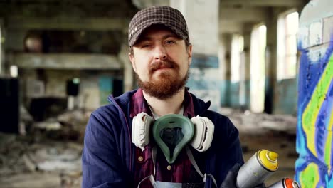 Close-up-portrait-of-handsome-bearded-man-graffiti-artist-standing-inside-abandoned-building-wearing-cap,-gloves-and-pespirator-and-holding-spray-paint