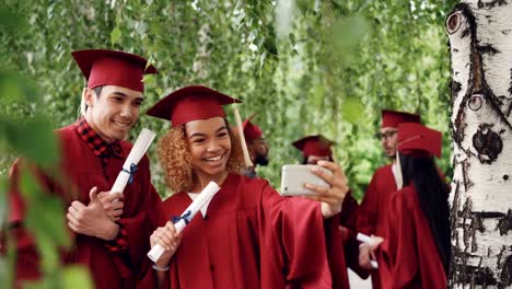 Happy-young-people-girl-and-guy-are-taking-selfie-after-graduation-ceremony-holding-diplomas-wearing-gowns-and-mortarboards.-Photographs-and-education-concept.