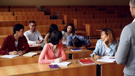 Tired-mixed-race-student-is-sleeping-during-lecture-at-college,-his-classmates-are-waking-him-up,-he-is-standing-and-talking-to-teacher,-young-people-are-laughing.