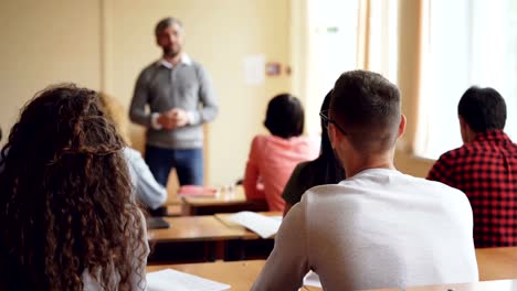 Participant-of-youth-conference-young-man-in-glasses-is-raising-hand-and-talking-to-tutor-while-other-people-are-looking-at-educator-and-listening-to-conversation.