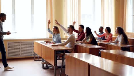 Young-people-students-are-raising-hands-and-answering-teacher's-questions-while-bearded-man-professor-is-speaking-and-gesturing.-Friendly-atmosphere,-light-room.