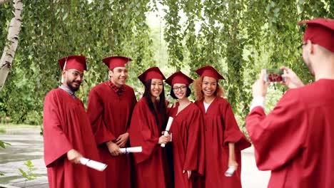 Multiethnic-group-of-graduating-students-is-posing-for-smartphone-camera,-waving-hands-with-diplomas-and-shouting-while-guy-is-shooting-them-touching-screen.