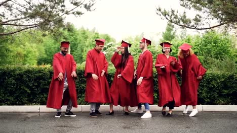 Emotional-young-people-in-mortar-boards-and-gowns-are-dancing-and-laughing-having-fun-with-green-trees-and-nature-in-background.-Education-and-celebration-concept.