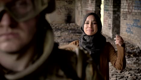 Young-muslim-woman-in-hijab-screaming-and-crying-behind-armed-soldier,-standing-in-abandoned-building,-war-concept