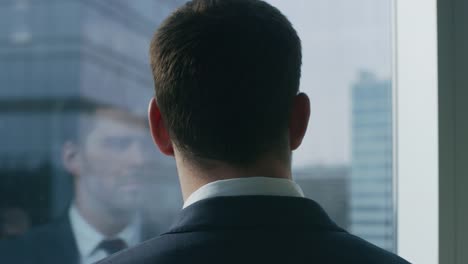 Close-up-Shot-of-the-Thoughtful-Businessman-in-a-Suit-Standing-in-His-Office-and-Contemplating-Next-Big-Business-Deal,-Looking-out-of-the-Window.-Big-City-Business-District-Panoramic-Window-View.