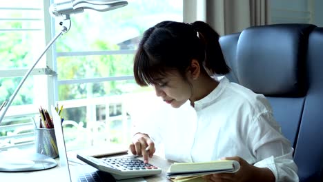 Student-learning-online-study-concept:-Beautiful-Asian-girl-listening-with-Headphones-and-laptop,-sitting-smile-writing-notes-in-textbook-at-her-desk-in-home-for-e-learning-in-educational-technology