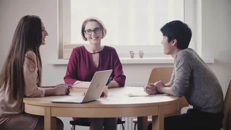 Zwei-schöne-positive-Kaukasische-Mädchen-sprechen-Sie-mit-japanischen-Mann-bei-einem-Vorstellungsgespräch-am-Tisch-in-moderne-gesunde-Büro