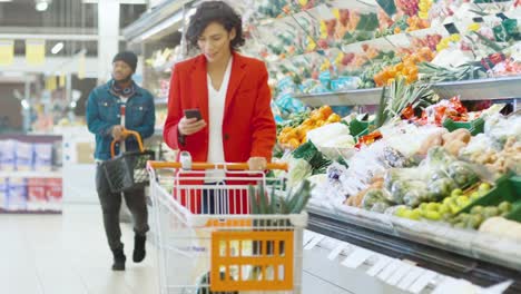 Im-Supermarkt:-schöne-junge-Frau-mit-Warenkorb-nutzt-Smartphone-und-Spaziergänge-durch-frisch-produzieren-Abschnitt-des-Speichers.