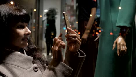 Shopping-at-mall,-sales-time.-Woman-looking-at-boutique-showcase-in-evening-city-on-street-and-takes-a-photo.-Woman-near-shop-window-with-mannequins.-Reading-bar-codes-by-smartphone.