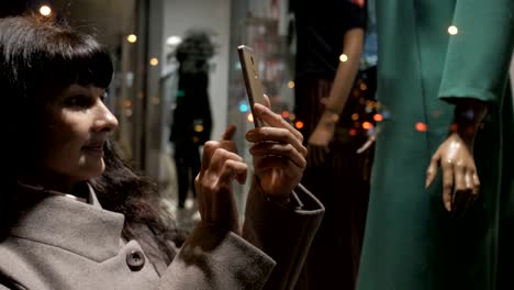 Compras-en-centro-comercial,-tiempo-de-ventas.-Mujer-mirando-el-escaparate-de-la-boutique-en-la-ciudad-de-noche-en-la-calle-y-toma-una-foto.-Mujer-cerca-de-escaparate-con-maniquíes.-Lectura-de-códigos-de-barras-por-smartphone.