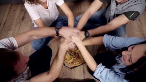Top-view-of-young-people-taking-hand-to-hand-under-the-box-with-pizza-then-take-slices-of-hot-tasty-pizza-from-cardboard-box.-Siting-on-the-wooden-floor,-drinks-and-pizza-on-the-floor